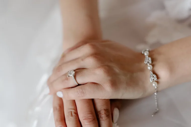 Bride's hands resting on a white lace dress, with white nails, a ring and a bracelet