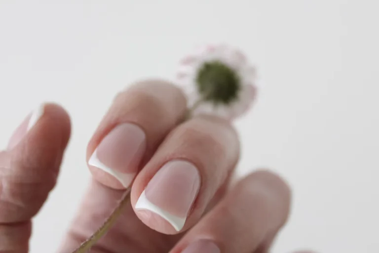 Hand with a classic French manicure holding a small daisy against a white background