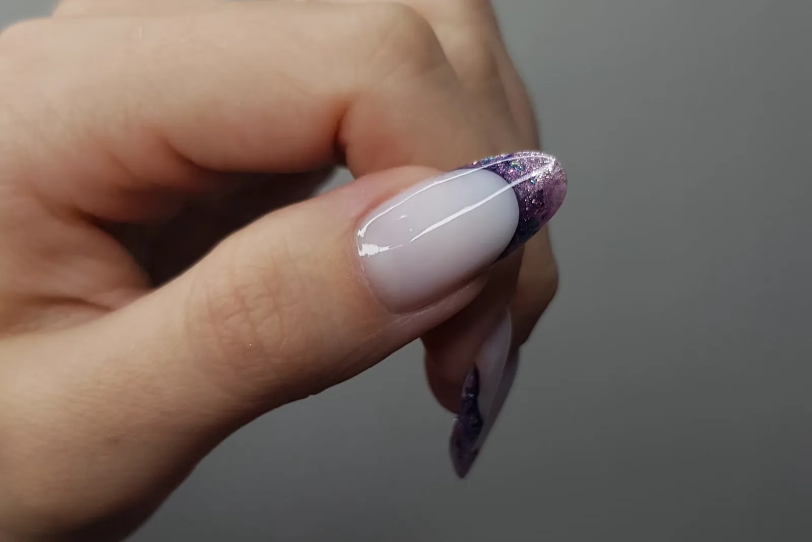 Hand with a pale lilac almond nail and a purple glitter French tip against a grey background