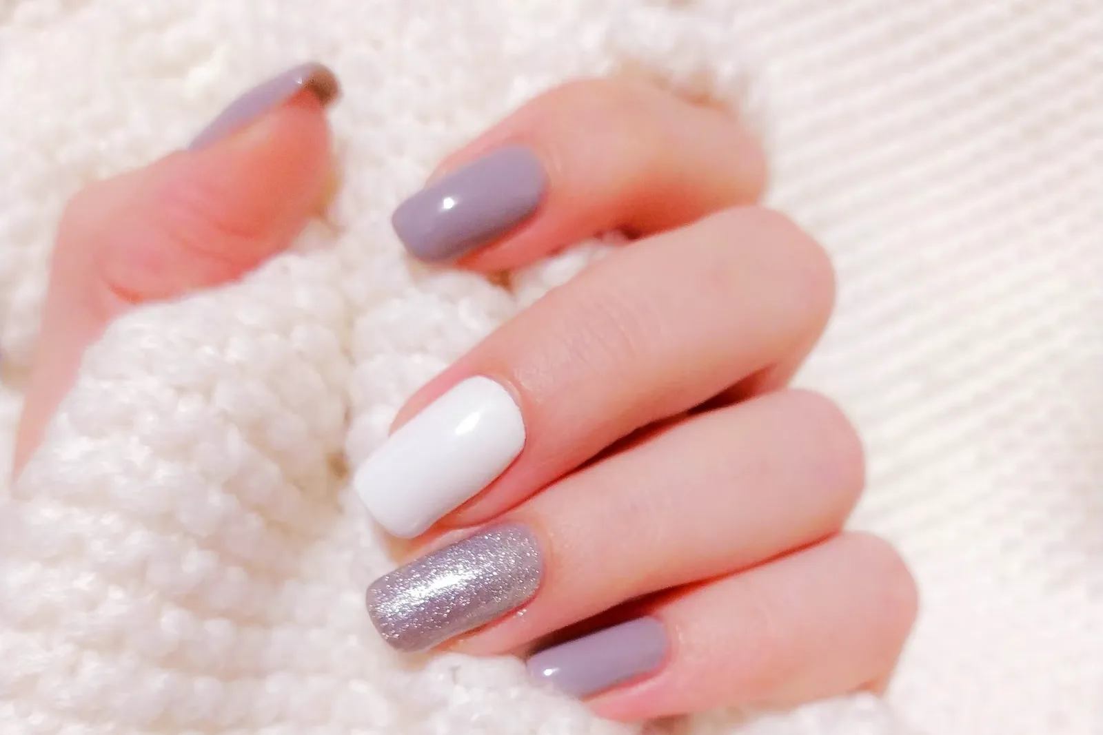 Hand with grey mauve, white and silver glitter nails resting on white knit fabric