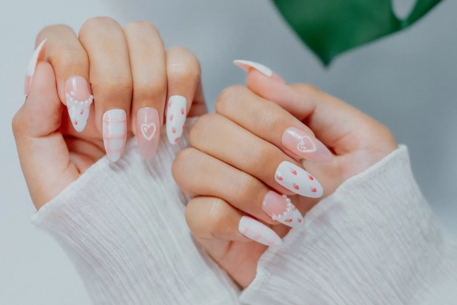 Two hands in white sleeves with pale pink and white nails decorated with hearts and dots beside a monstera leaf